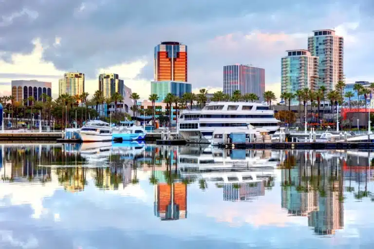 Scenic waterfront view of downtown Long Beach, California, featuring modern high-rise buildings, palm trees, and luxury yachts docked in the marina with clear reflections on the calm water under a partly cloudy sky.