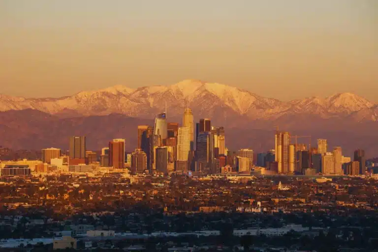Downtown Los Angeles skyline with snow-capped San Gabriel Mountains glowing at sunset, viewed from the South Gate area.