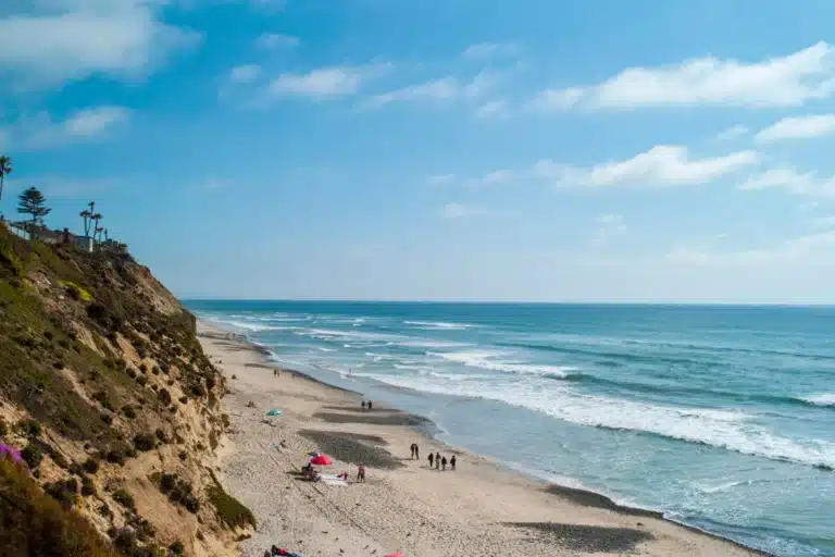 Wide view of Encinitas beach showing ocean waves, sandy shoreline, and coastal cliffs under blue sky.