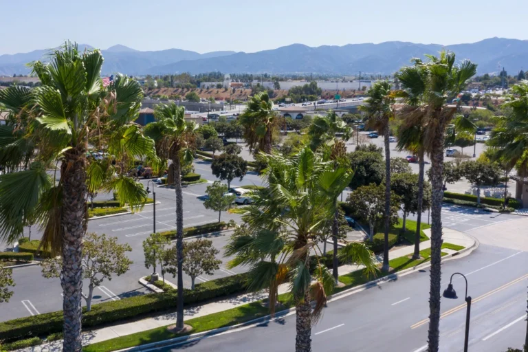 Aerial view of a Corona, California neighborhood with a church, residential streets, palm trees, and cityscape stretching toward distant mountains.